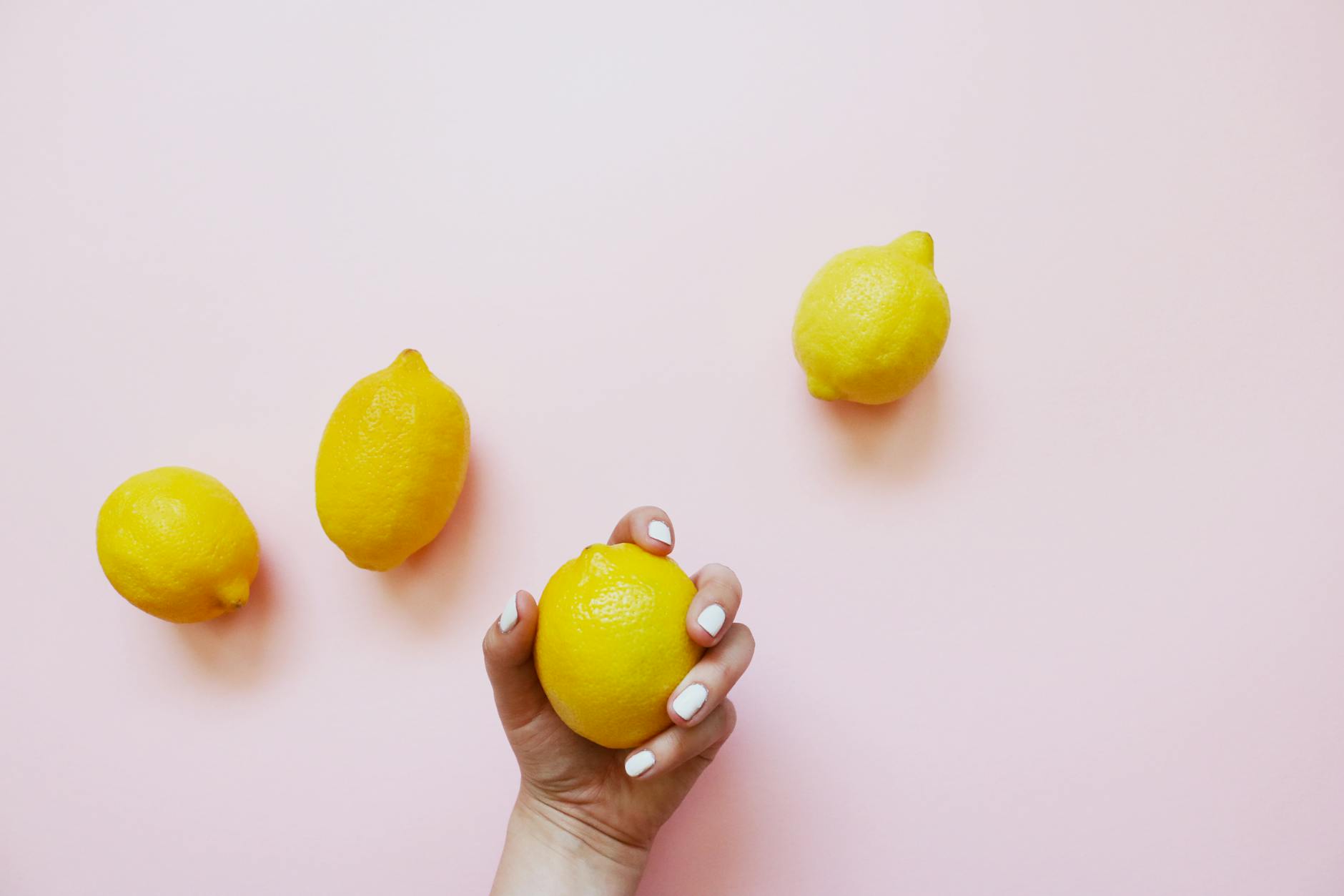 A hand holding a fresh lemon on a soft pink background with additional lemons nearby, symbolizing renewed sensation and pleasure